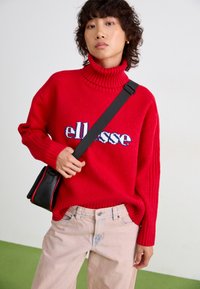 Sarkans augstais kakls džemperis ar ribotu tekstūru un izteiksmīgu "ellesse" logo zilā un baltā krāsā, apvienots ar gaiši rozā biksēm un melnu plecu somu.