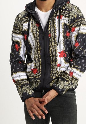 Mężczyzna w czarno-biało-złotej bluzie z zamkiem błyskawicznym i wzorem, ozdobionej czerwonymi różami, noszący białą koszulkę i czarne dżinsy.