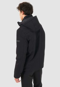 Giacca outdoor nera con cappuccio, realizzata in un tessuto liscio e opaco e con un logo discreto sulla spalla sinistra. Design semplice con una vestibilità sartoriale.