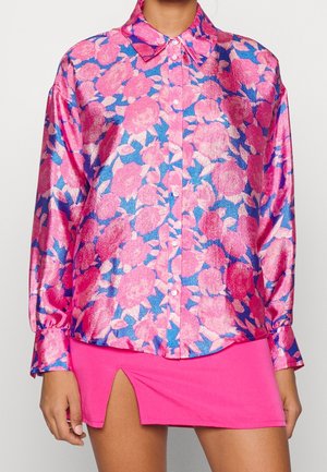 Camisa de manga larga con botones y estampado floral brillante en rosa y azul, combinada con minifalda rosa fuerte con abertura frontal.