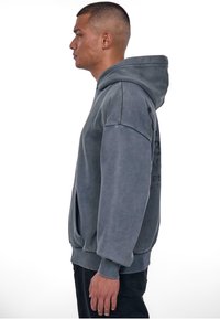 Grau, übergroßer Kapuzenpullover mit einer Fronttasche, gerippten Bündchen und matter Oberfläche. Besondere Merkmale sind ein grafischer Druck auf der Rückseite.