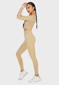 Beige langärmliger Cropped Pullover mit weißen Streifen an den Ärmeln und hochgeschnittenen Leggings. Glatte Textur und figurbetontes Design. Weißes Logo-Detail.