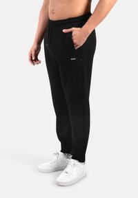 Muž v černých teplácích se stojícími rukama v kapsách a bílými teniskami, stojící na jednobarevném světlém pozadí.