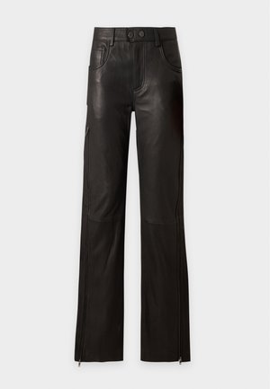 Femme portant une chemise blanche boutonnée et un pantalon en cuir noir à jambes larges, tenant une pochette noire froncée sur fond blanc.