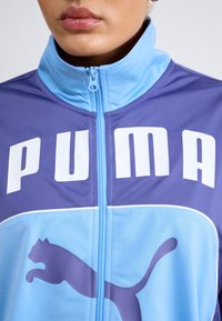 Person som bär en blå och lila zip-up jacka med stor vit "PUMA"-text och en lila puma-logotyp på framsidan.