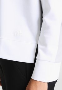 Sudadera blanca de manga larga, con puños acanalados y un logo sutil. Tejido suave con una textura lisa, combinada con pantalones negros.