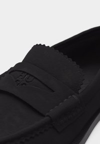 Μαύρο μοκασίνι από σουέντ με κυματιστές άκρες, λεπτομέρεια ραφής μοκασίνου και διακοσμητικό λουρί στο επάνω μέρος.