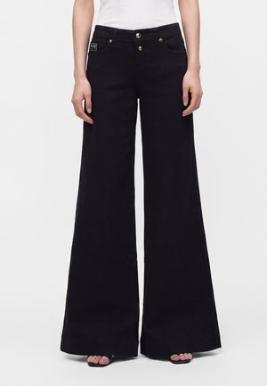 Femme portant un pantalon noir taille haute à jambes larges avec des boutons dorés et un haut blanc, debout devant un fond clair uni.