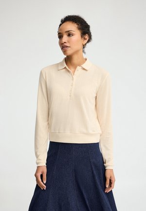 Kobieta o kręconych włosach, ubrana w kremową koszulkę polo z długim rękawem i ciemnoniebieską rozkloszowaną spódnicę, stoi na tle jednolitego jasnego tła.