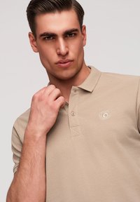 Homme ajustant le col d’un polo beige avec logo brodé sur fond uni clair.