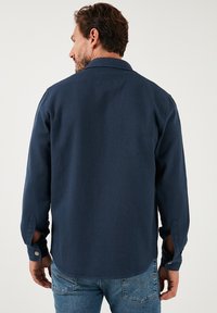 Camicia a maniche lunghe blu navy in tessuto strutturato, con colletto e polsini con bottoni. La vista posteriore mostra un orlo dritto e cuciture dettagliate.
