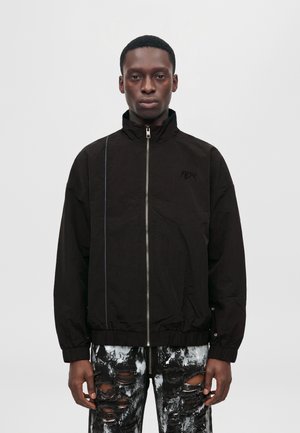 Un mannequin se tient devant un fond uni, vêtu d’une veste noire zippée et d’un jean noir et blanc à l’aspect usé.