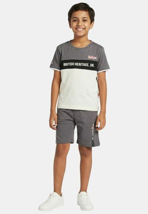 Garçon debout, souriant, portant un t-shirt gris et blanc "British Heritage. UK.", un short gris assorti et des baskets blanches.