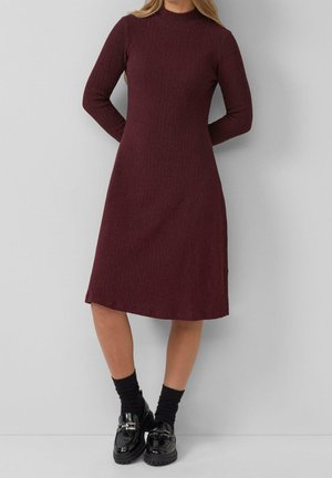 Femme portant une robe bordeaux côtelée à manches longues et longueur genou, avec des chaussettes noires et des mocassins noirs brillants, debout contre un mur gris clair uni.