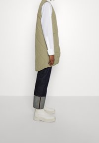 Gequilted olijfgroene vest, oversized ontwerp, gecombineerd met een witte blouse met kraag. Donkerdenim broek met opgerolde pijpen en ritsdetails. Stevige witte laarzen.