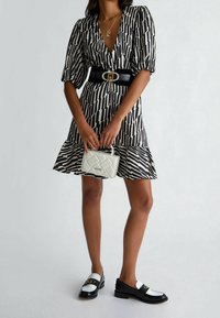 Frau in einem schwarz-weißen gemusterten Kleid mit Puffärmeln, schwarzem Gürtel, die eine kleine weiße gesteppte Handtasche und schwarz-weiße Slipper hält.
