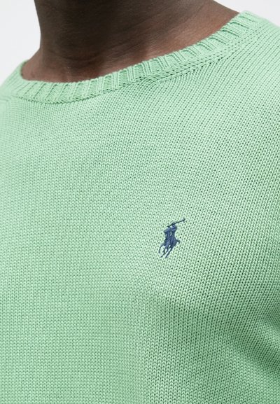 Svetlo zelena pletena jopica z rebrastim ovratnikom in majhnim mornarsko modrim logotipom na levem prsnem delu. Enostavna tekstura po celotni površini.