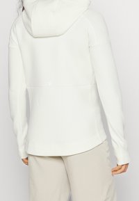 Vit huva sweatshirt med en slät textur, som har en figursydd design, lång ärm och en platt söm på baksidan. Diskret logotypdetalj under huvan.