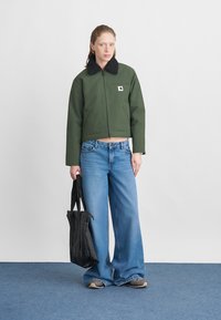 Geknipte groene jas met zwarte fleece kraag, gecombineerd met een wijdvallende blauwe spijkerbroek. Model houdt een zwarte totesac vast en staat op een blauwe ondergrond.