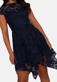 Navy blue dress with sheer floral lace overlay, flared skirt with asymmetrical hem, cap sleeves, and intricate embroidery details.