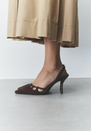 Stopa kobiety noszącej brązowy zamszowy sandał na wysokim obcasie typu slingback z wycięciami, stojącej na szarej podłodze, widoczny beżowy brzeg spódnicy nad kostką.