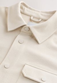 Chemise en tissu texturé écru avec un col pointu, des boutons-pression sur le devant et une poche à rabat boutonnée sur la poitrine.