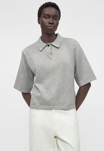 Άτομο που φοράει φαρδιά γκρι πλεκτή μπλούζα πόλο με κουμπιά και φαρδιά μανίκια, σε συνδυασμό με ψηλόμεσο λευκό παντελόνι, που στέκεται μπροστά σε απλό φόντο.