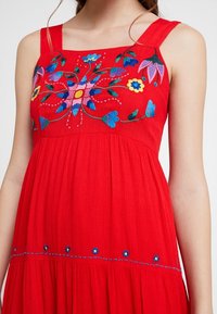 Robe rouge sans manches ornée d'une broderie florale vive en bleu, rose et jaune, avec une jupe plissée et des coutures décoratives le long de l'ourlet.