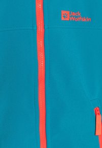 Giacca softshell verde acqua con una cerniera e dettagli arancioni vivaci. Presenta un piccolo logo e una tasca laterale con una cerniera a contrasto.