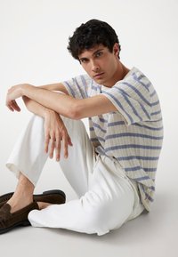 Top de punto de manga corta a rayas en crema y azul claro. Combinado con pantalones blancos y mocasines de ante marrón. Modelo sentado, con los brazos descansando sobre las rodillas.