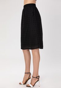 Jupe noire mi-longue avec un design en œillets texturés, taille élastique et petites découpes décoratives le long de l'ourlet. Assortie à des chaussures à talons blanches et noires.