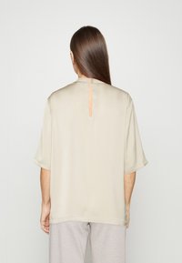 Beige kortærmet top med rund halsudskæring, der har en nøglehulåbning i ryggen og en glat, blød tekstur. Enkel, ude ornamenteret design.