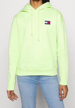 Personne portant un sweat à capuche Tommy Jeans vert clair avec poche avant et un jean bleu clair sur un fond blanc uni.