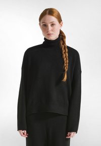 Schwarzer Rollkragenpullover aus weichem Material, mit entspanntem Schnitt, überschnittenen Schultern und geripptem Saum, kombiniert mit passenden schwarzen Unterteilen.