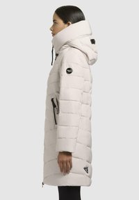 Light grey puffer coat with a high collar, quilted design, side zip pockets, and logo patch on the arm. Features a drawstring hood.