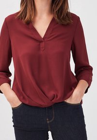 Bordeauxfarvet bluse med V-hals og 3/4 ærmer, lavet af et glat stof. Har en knuddet detalje ved taljen og en afslappet pasform.
