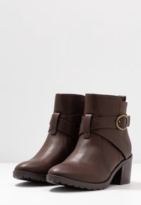 Botas de tobillo de cuero marrón con puntera redondeada, una correa decorativa y un detalle de hebilla, y un tacón apilado con una suela texturizada.