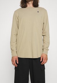 Uomo in camicia a maniche lunghe beige con un piccolo logo sul petto e pantaloni cargo neri, in piedi contro uno sfondo bianco.