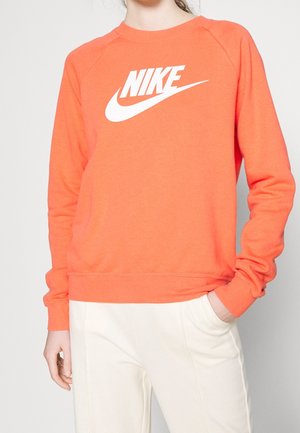 Personne portant un sweat-shirt orange Nike avec un logo blanc et un pantalon couleur crème, posant avec une main dans la poche et l’autre pendant.