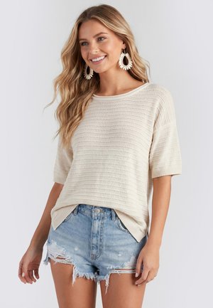 Lachende vrouw met lang golvend blond haar, grote witte oorbellen, een crèmegekleurde gebreide top met korte mouwen en versleten lichtblauwe spijkershorts.