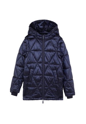 Kind trägt dunkelblaue gesteppte Jacke, Colorblock-Pullover, weit geschnittene Jeans und weiße Sneakers, steht neben Pinseln und Papier auf dem Boden.