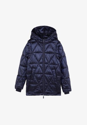 Enfant portant une veste matelassée bleu marine, un pull color block, un jean à jambes larges et des baskets blanches, debout à côté de pinceaux et de papier sur le sol.