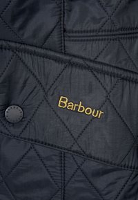Pikowana ciemnogranatowa kurtka z okrągłym czarnym guzikiem i złotym logo "Barbour". Gładka faktura z detalami w kształcie diamentów.