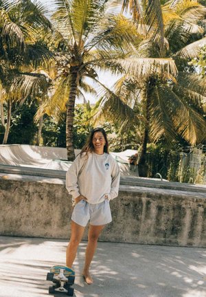 Jasnoszara bluza, paskowane niebiesko-białe spodenki, boso. Stoi obok deskorolki na betonowej powierzchni, otoczony palmami.