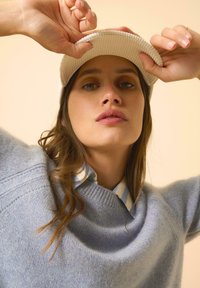 Lichtblauwe gebreide trui met een losse halslijn, gedragen over een lichtblauw en wit gestreept shirt, vergezeld van een crèmekleurige cord cap.