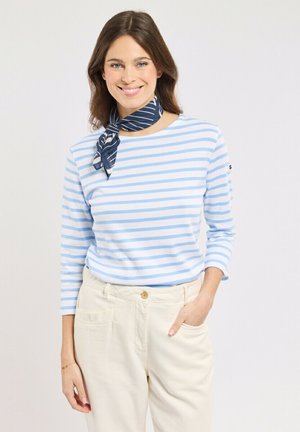 Femme souriante, portant une chemise à manches longues à rayures bleu clair et blanches, un pantalon beige, un foulard à rayures bleu marine autour du cou, une main dans la poche.