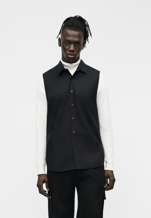 Mann mit Dreadlocks, der ein schwarzes, ärmelloses Hemd über einem weißen Langarm-Rollkragenpullover und schwarze Hosen vor einem schlichten Hintergrund trägt.