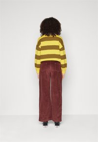 Geel en bruin gestreepte trui, gecombineerd met brede maroon corduroypantalons. Zwarte sneakers met witte accenten maken de look compleet.