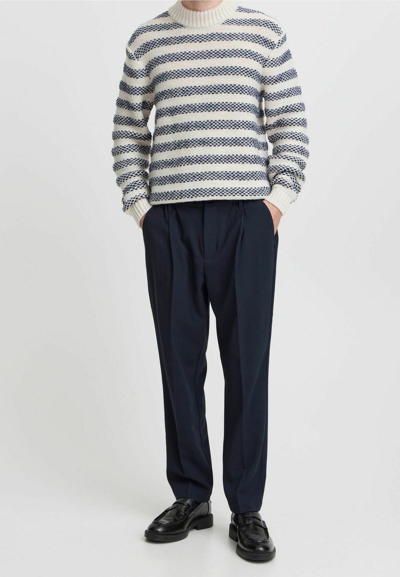 Homme portant un pull rayé texturé blanc et bleu marine, un pantalon plissé bleu marine, des mocassins en cuir noirs, debout les mains dans les poches devant un fond uni.