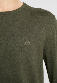 Lähikuva henkilöstä, joka wearing paksua oliivinvihreää neulepaitaa, jossa on pieni brodeerattu vuori- ja lippulogo rinnassa.